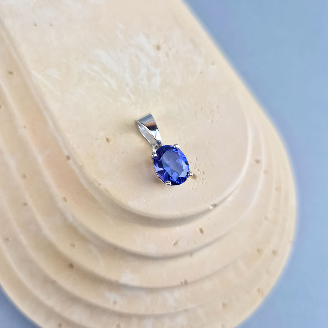 Melsvai violetinis tanzanito pakabukas sidabro tone ant smėlio spalvos padėklo su mėlynu fonu
