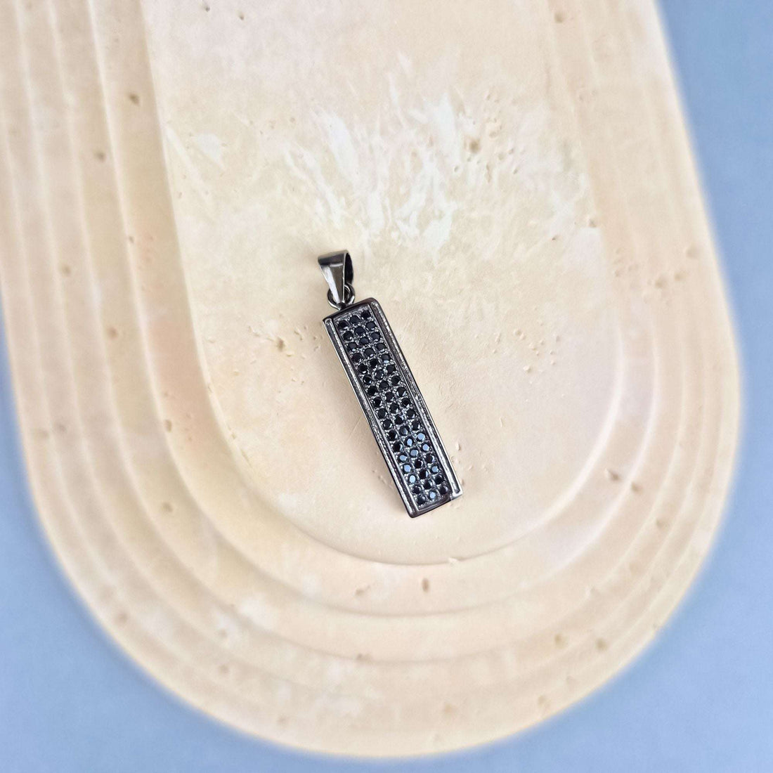 Pailgas juodų akmenukų pakabukas su kilpele, nufotografuotas įstrižu rakursu ant šviesaus akmens